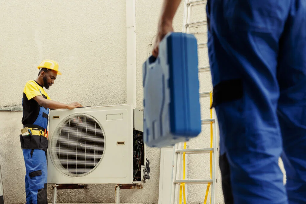 Two workers in uniforms and helmets service an outdoor air conditioning unit; one holds a blue toolbox while a ladder stands in the background. They also offer comprehensive air duct cleaning for improved efficiency.